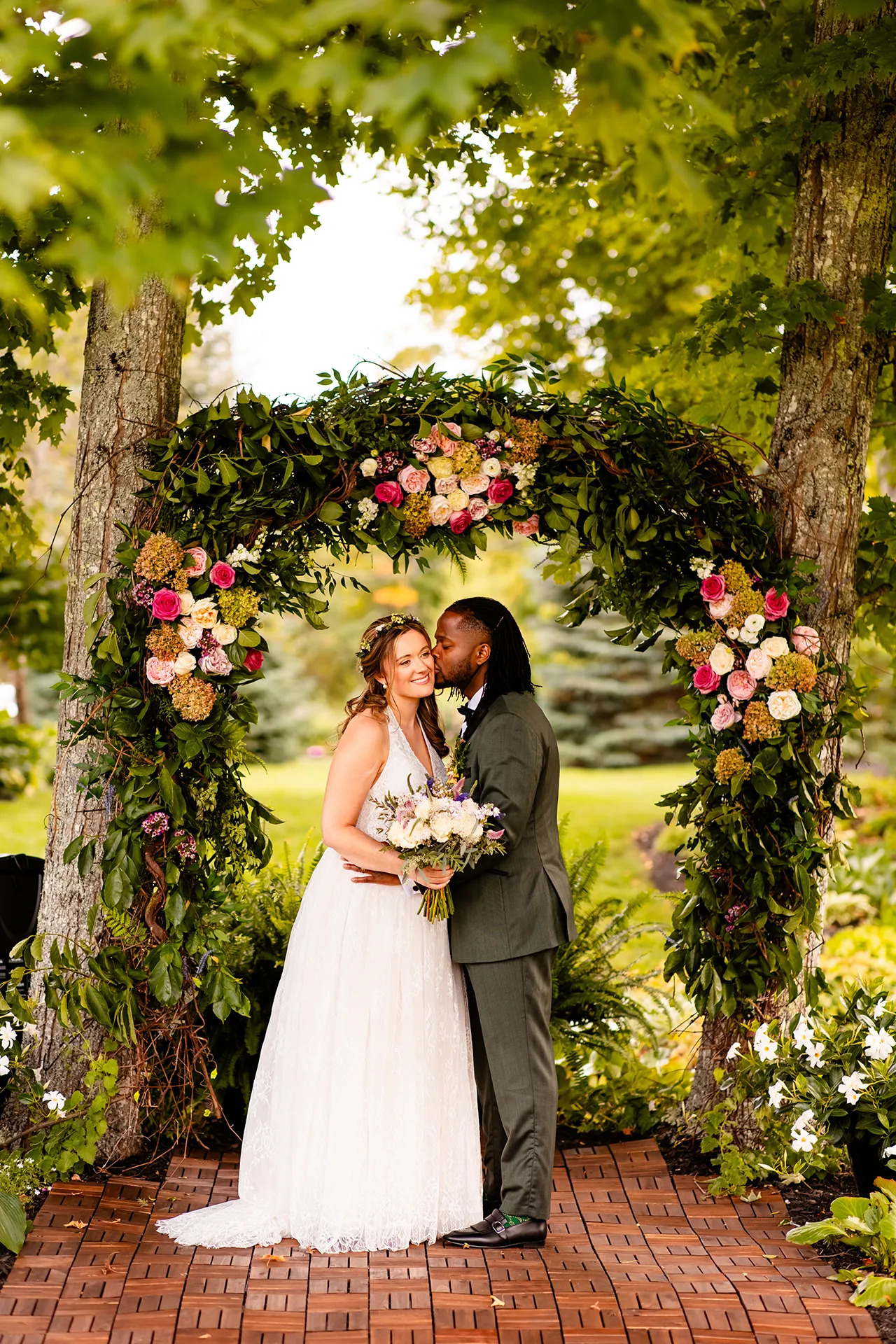 A bride smiles as a groom kisses her on the cheek in front of an arbor of flowers during wedding portraits in Bangor, Maine.