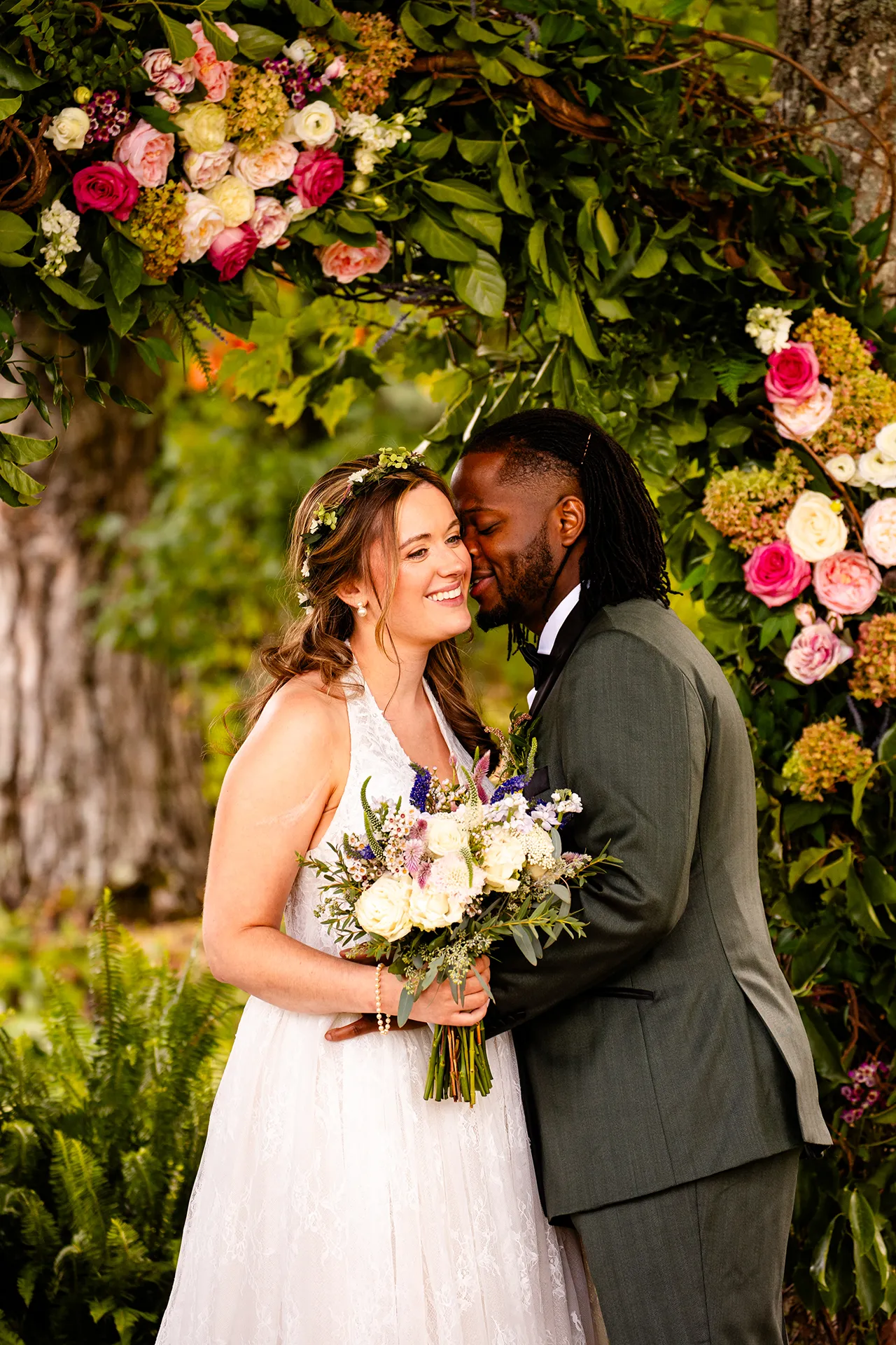A groom kisses a bride on the cheek while posing in front of a flower arbor during wedding portraits in Bangor, Maine.