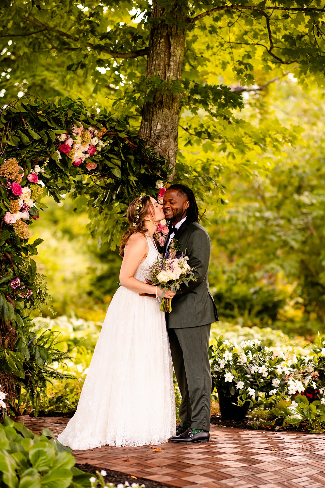 A bride kisses a smiling groom on the cheek in a flower garden during wedding portraits in Bangor, Maine.
