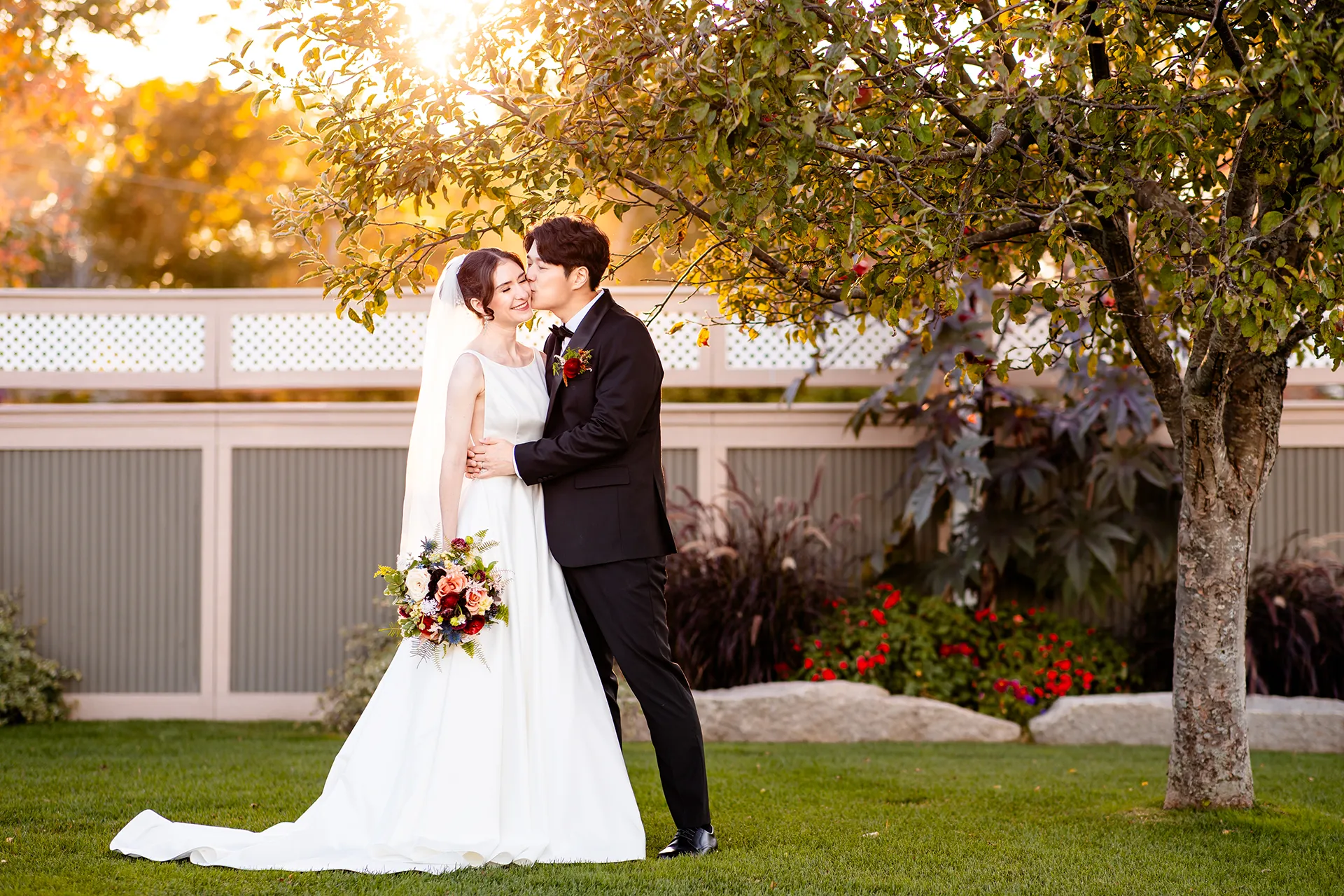A couple stands under a tree while a groom kisses a bride on the cheek at the Bar Harbor Club near Acadia National Park in Maine.