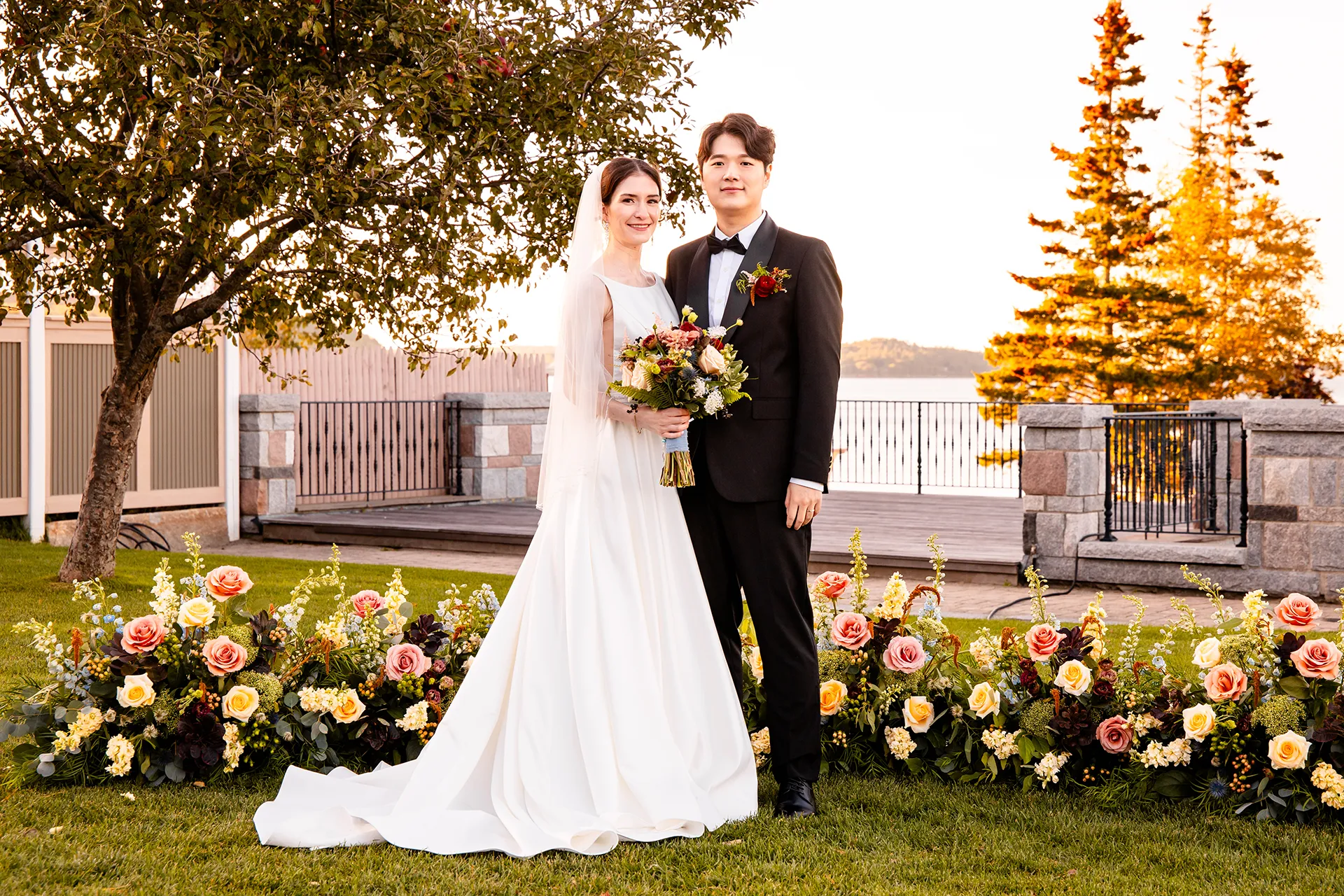 A bride and groom smiles and pose during wedding portraits in front of flowers at the Bar Harbor Club near Acadia National Park in Maine.
