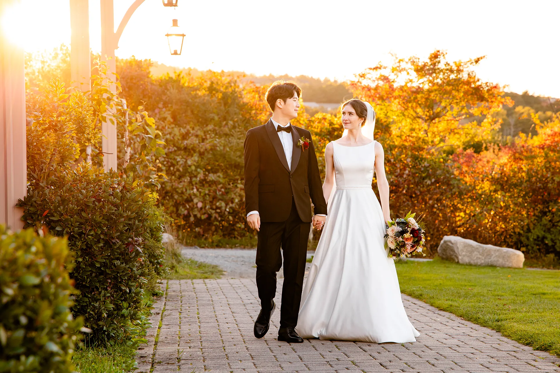 The sun shines behind a newly married couple as they hold hands and walk during portraits at the Bar Harbor Club near Acadia National Park in Maine.