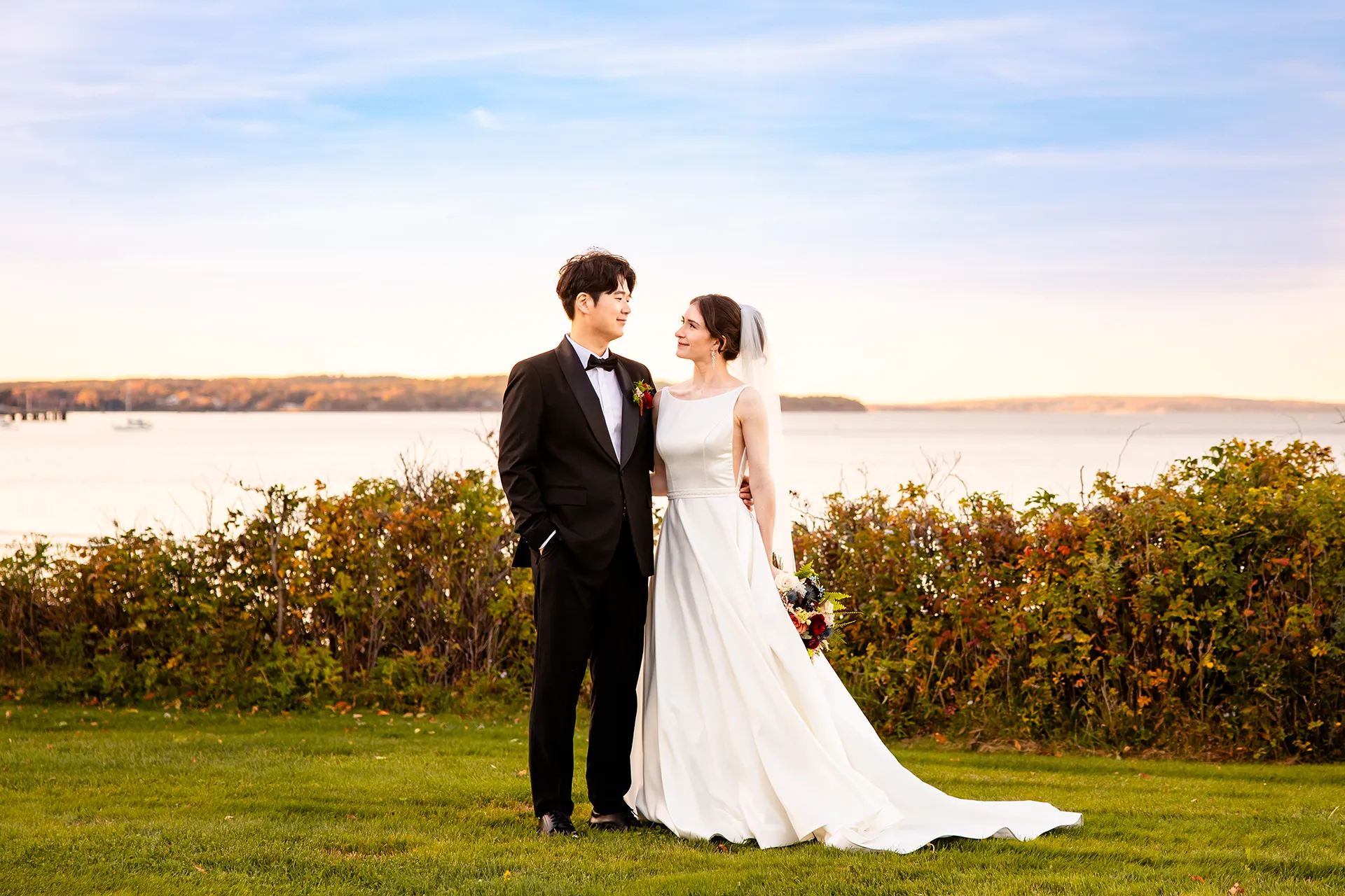 A bride and groom wrap their arms around each other in front of Frenchman Bay at the Bar Harbor Club near Acadia National Park in Maine.