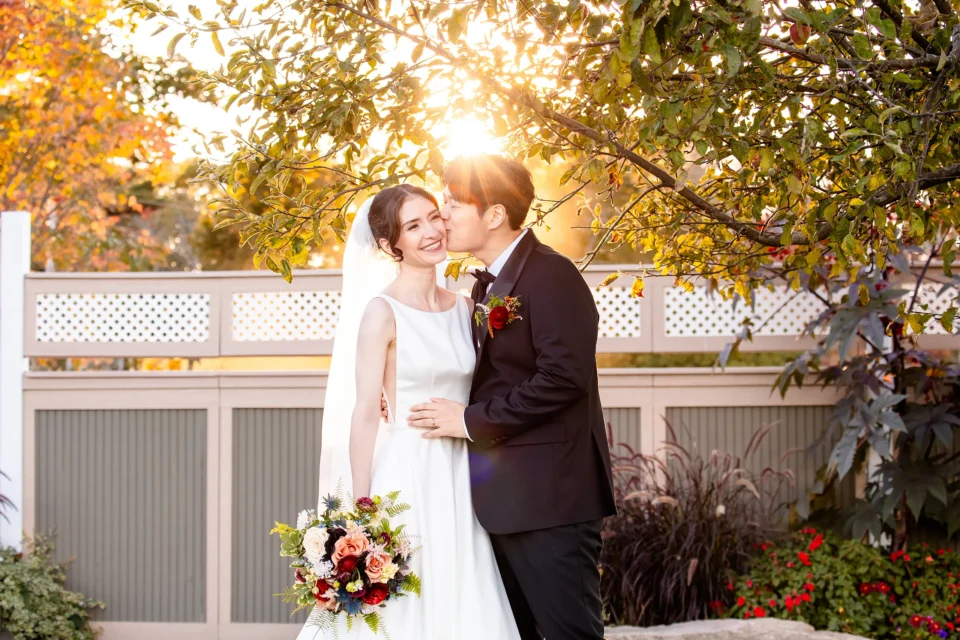 A bride smiles as a groom kisses her on the cheek as the sun shines behind them at the Bar Harbor Club near Acadia National Park in Maine.