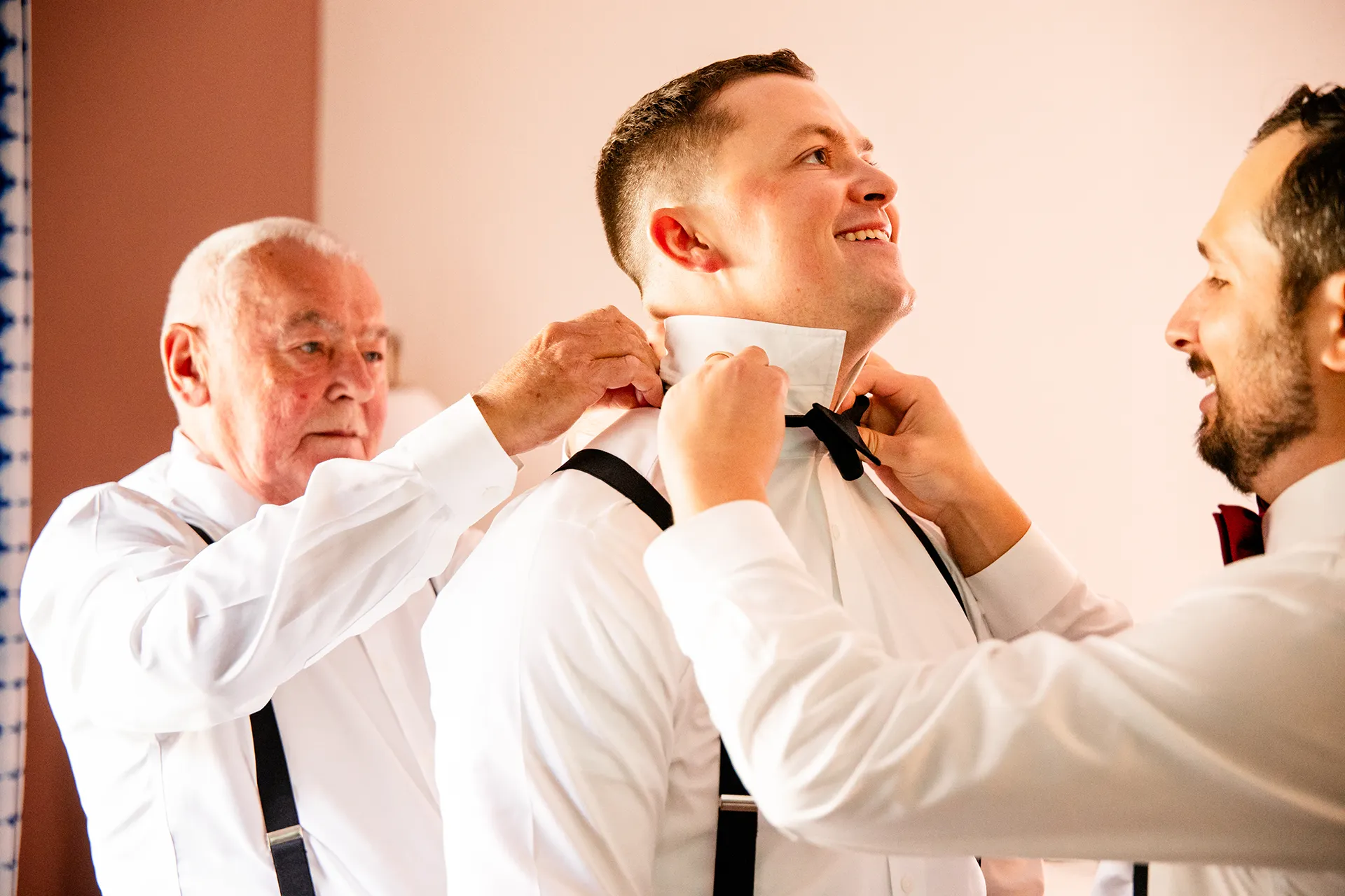 A dad and groomsman help a groom put on his bowtie as he gets ready for a wedding at Fruitlands Museum in Harvard Massachusetts.