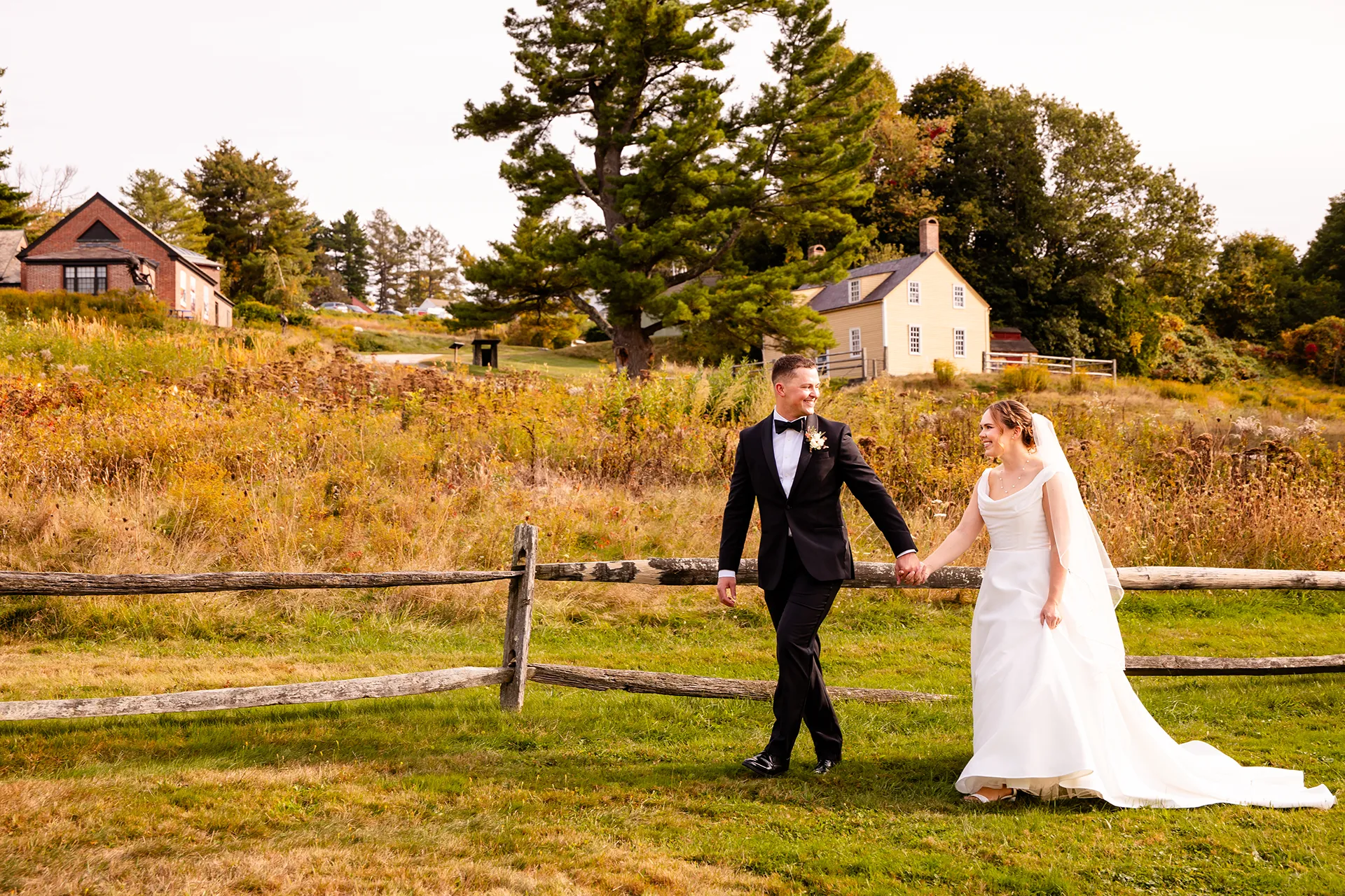 A bride and groom hold hands and walk in a field during wedding portraits at Fruitlands Museum in Harvard Massachusetts.