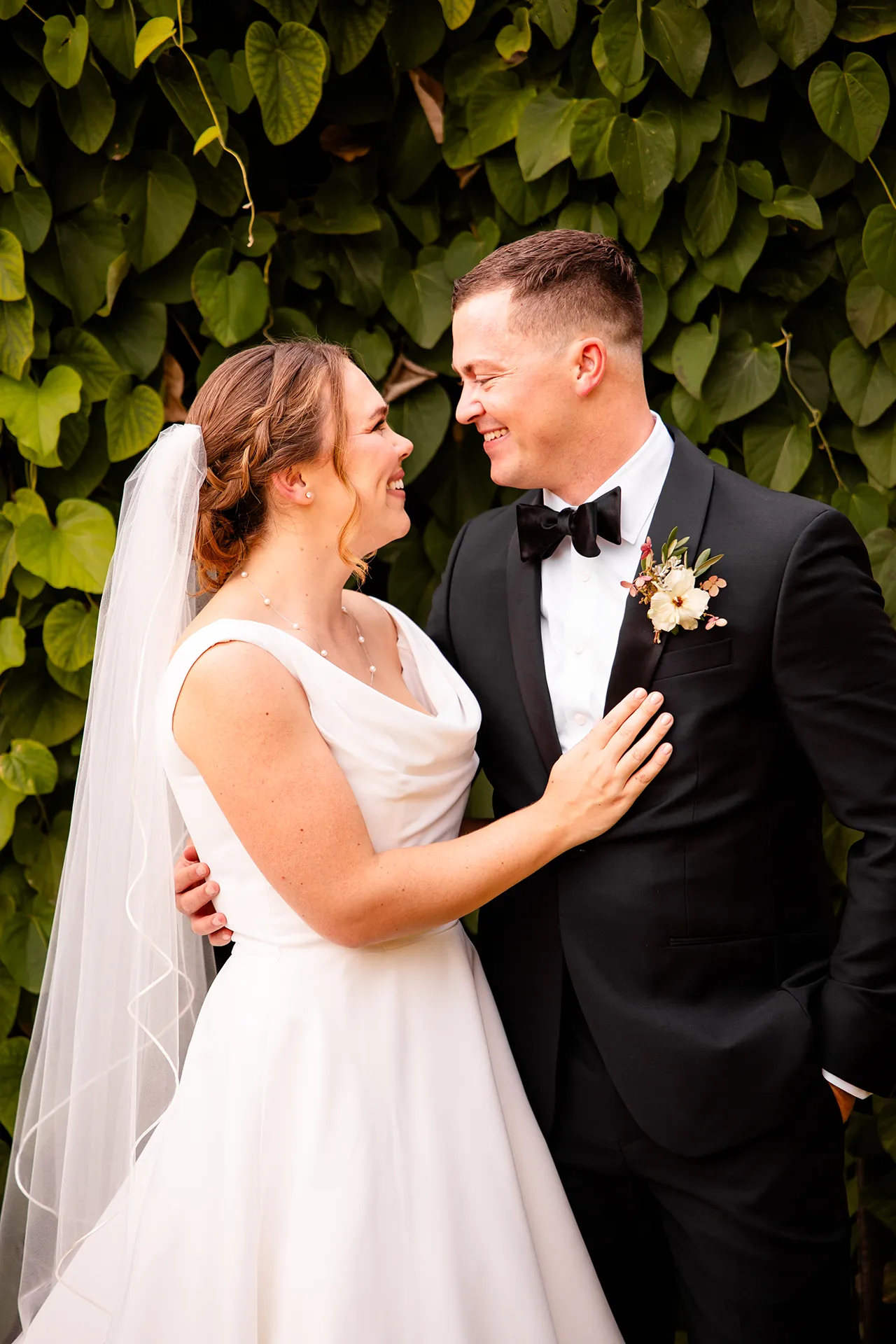 A bride and groom smile at each other in front of a wall of green leaves during wedding portraits at Fruitlands Museum in Harvard Massachusetts.