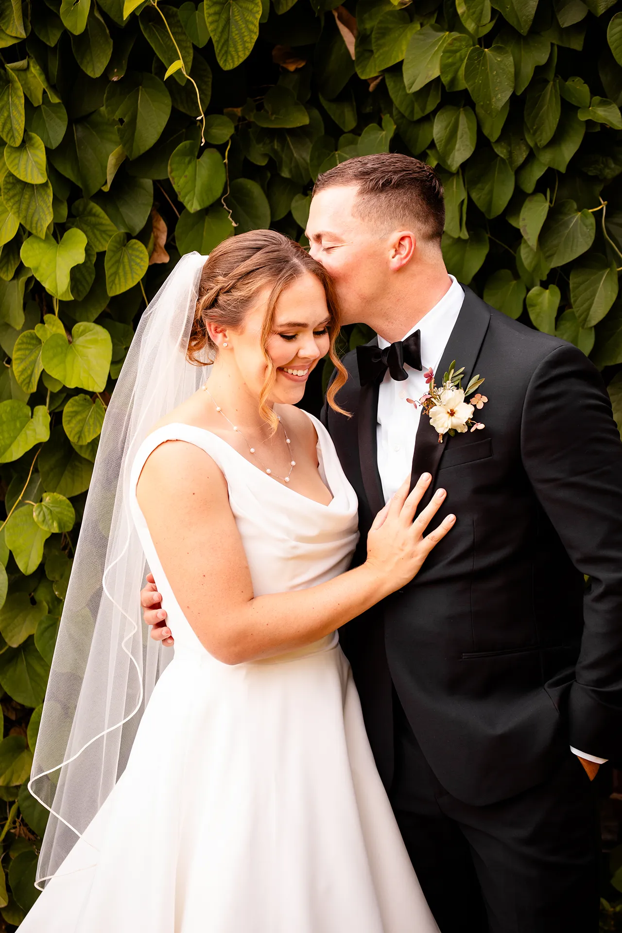 A groom kisses a bride on the head in front of a wall of green leaves during wedding portraits at Fruitlands Museum in Harvard Massachusetts.