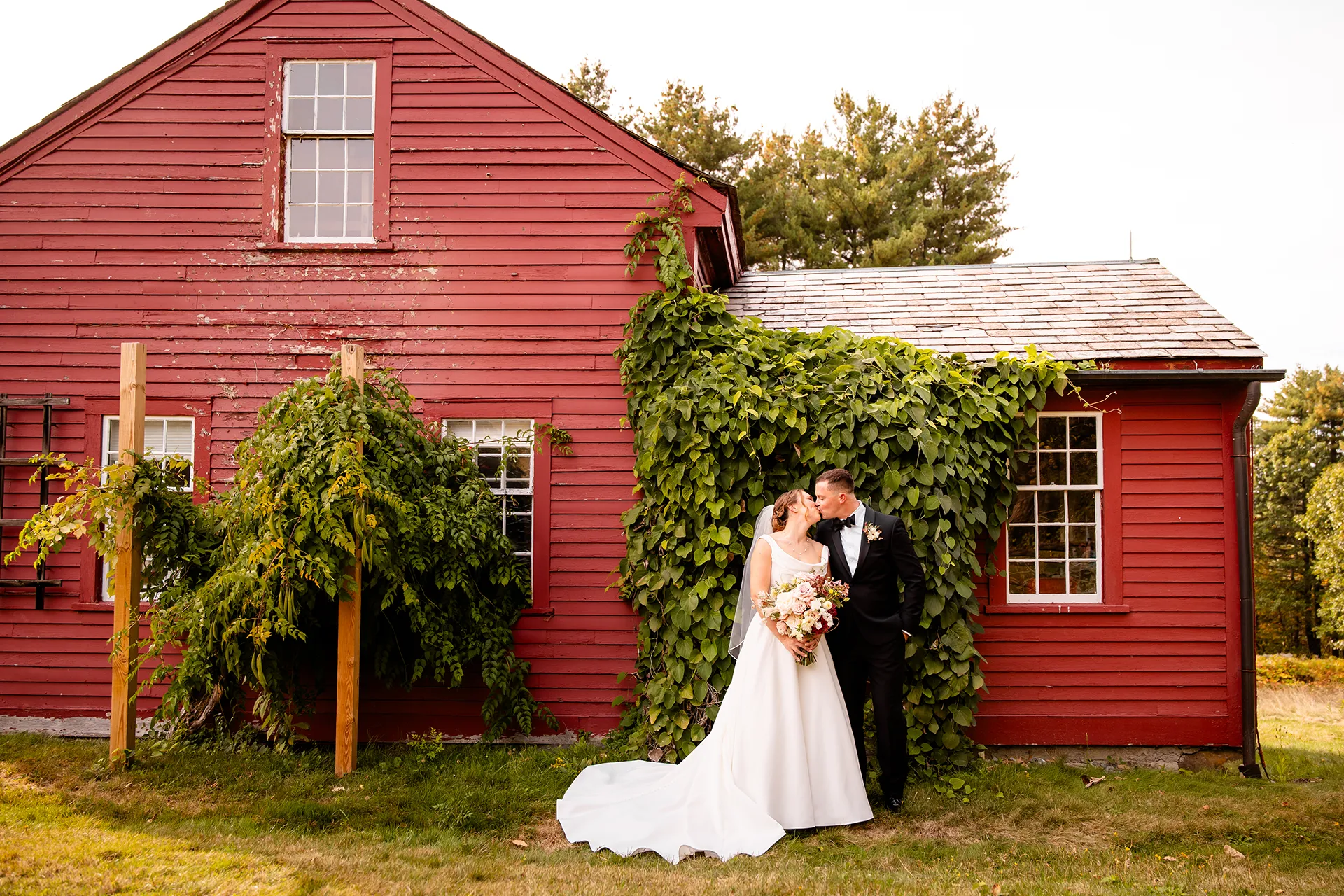 A bride and groom kiss in front of a red house during wedding portraits at Fruitlands Museum in Harvard Massachusetts.