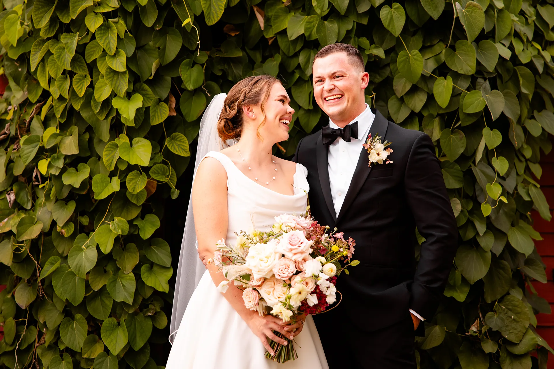 A bride and groom laugh while posing in front of a wall of green leaves during wedding portraits at Fruitlands Museum in Harvard Massachusetts.