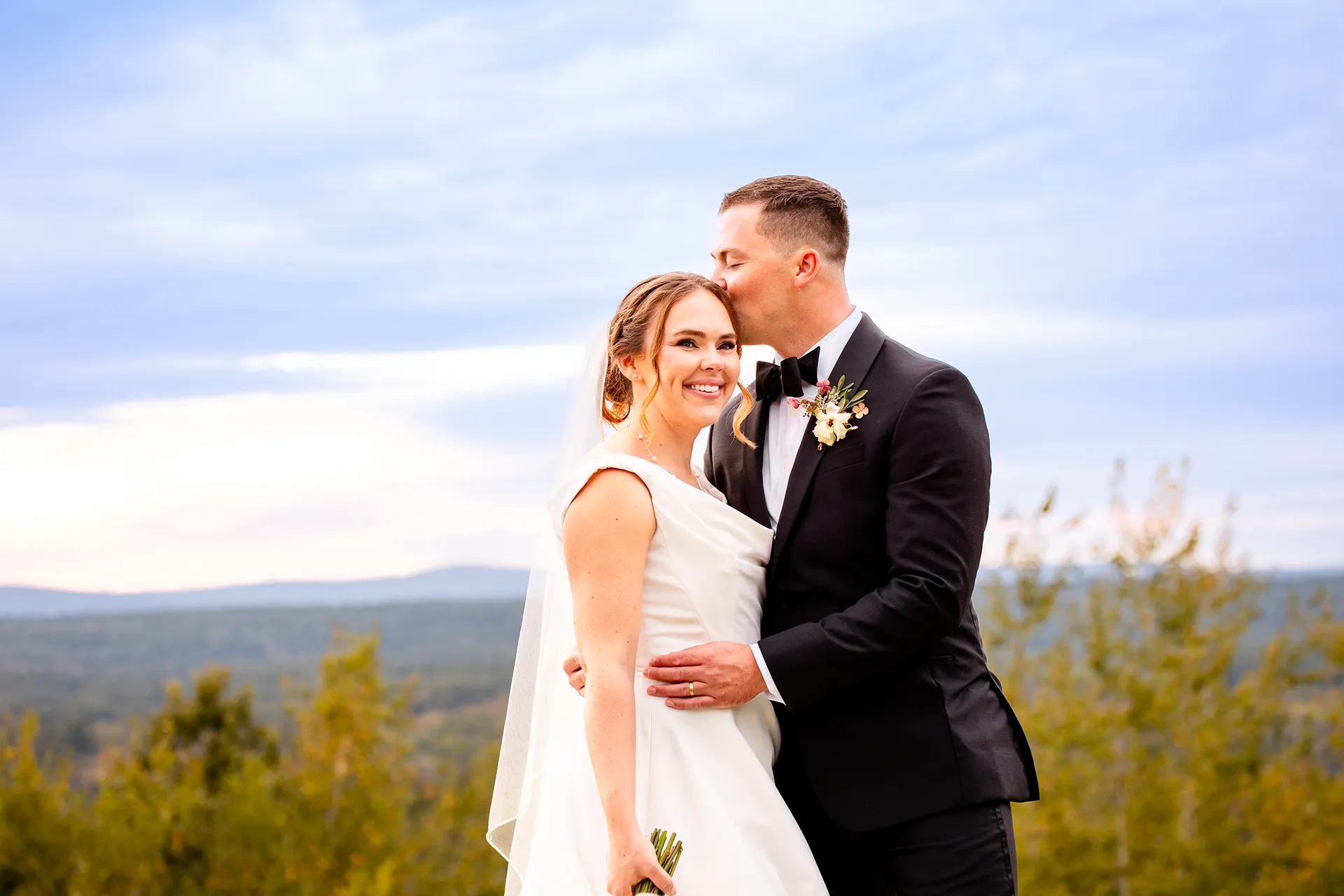 A bride smiles as a groom kisses her on the head during wedding portraits at Fruitlands Museum in Harvard, Massachusetts.