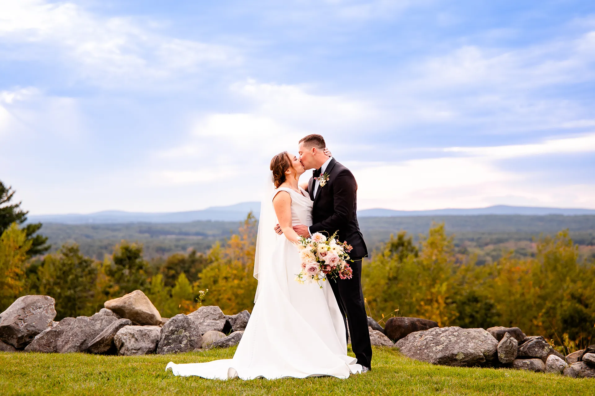 A bride and groom stand on a hill overlooking a mountain and kiss during wedding portraits at Fruitlands Museum in Harvard, Massachusetts.