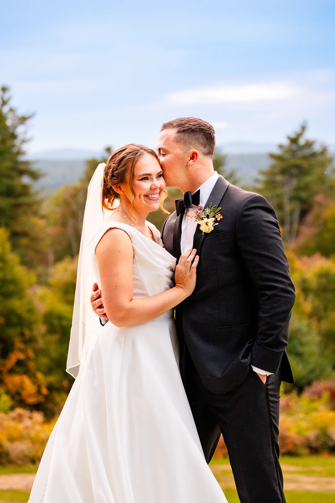 A bride smiles as a groom kisses her on the cheek during fall wedding portraits at Fruitlands Museum in Harvard, Massachusetts.