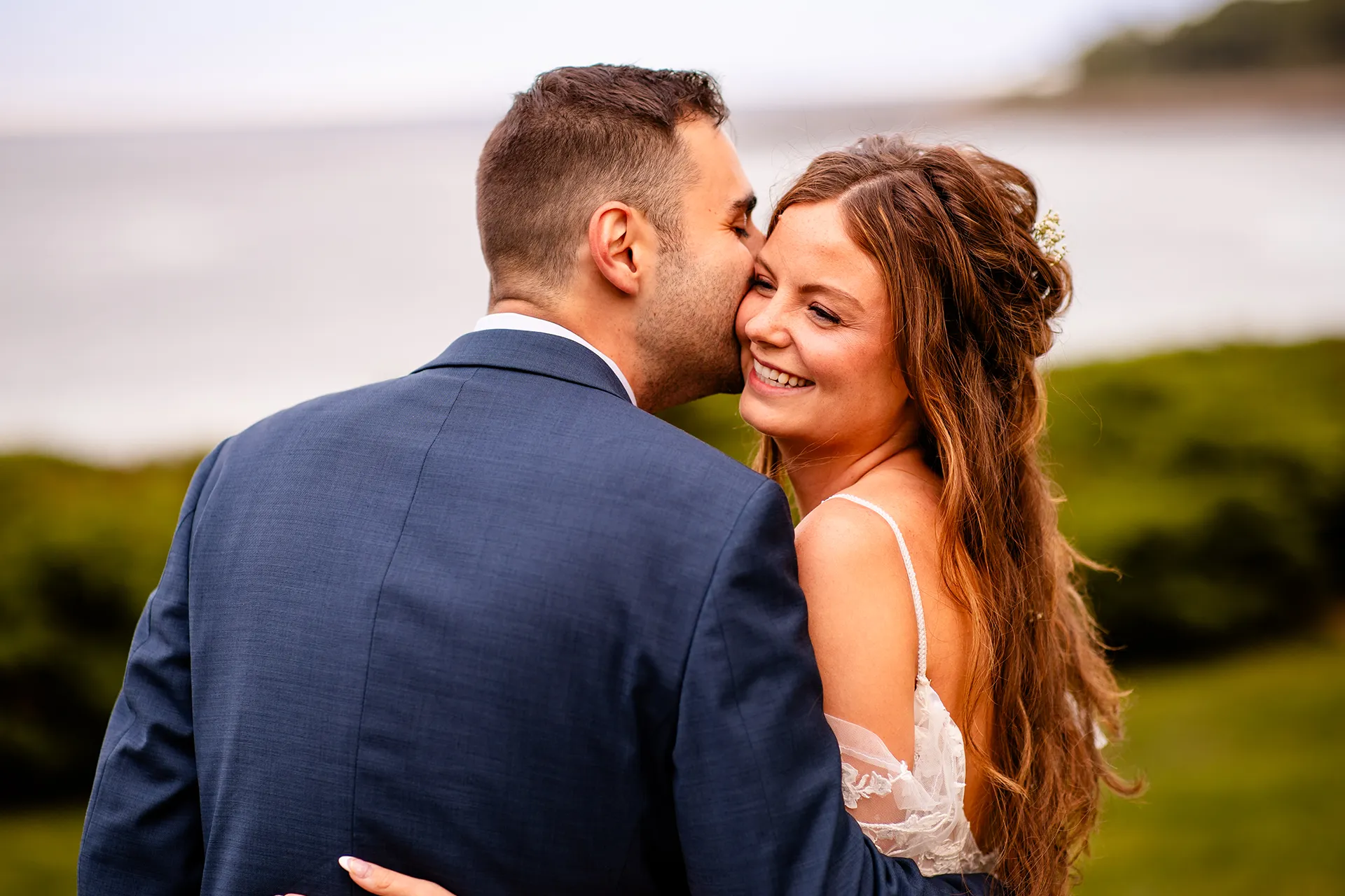 A bride smiles as a groom kisses her on the cheek during wedding portraits at the Hartley Mason Reserve near the York Harbor Reading Room in Maine.