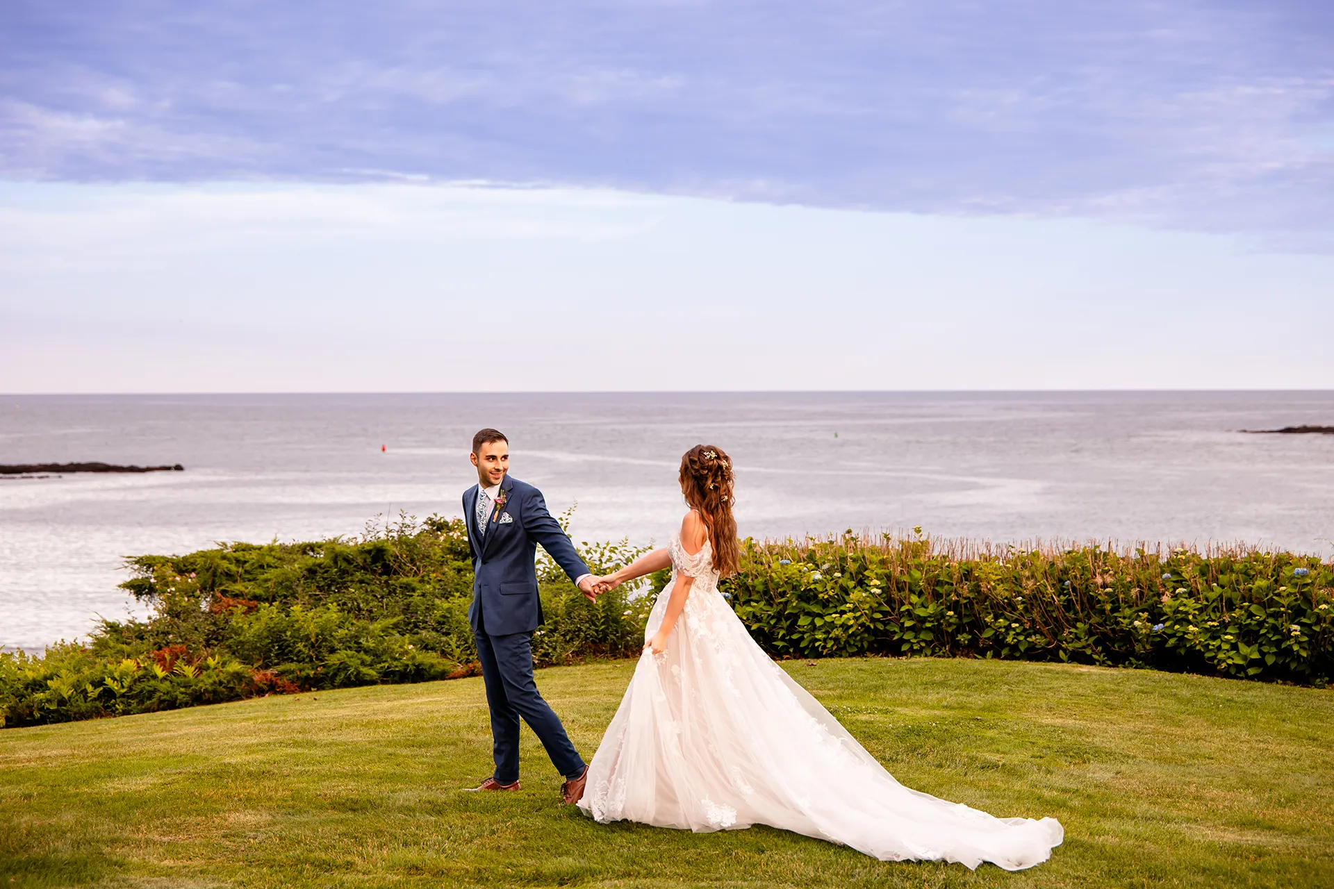 A groom looks back at a bride while walking during wedding portraits at Hartley Mason Reserve in York, Maine.