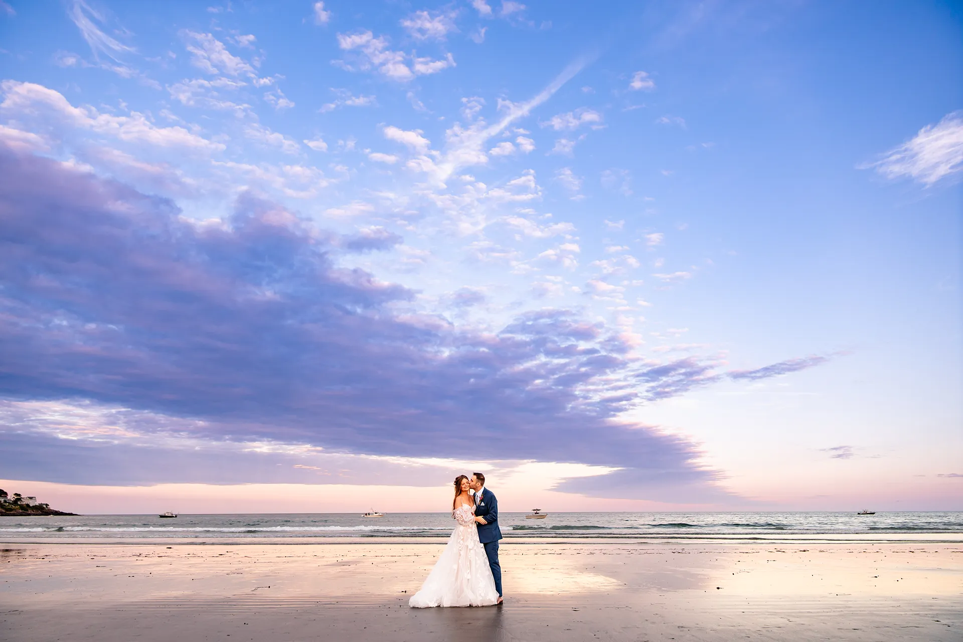 A bride smiles as a groom kisses her on the cheek during sunset wedding portraits at Long Sands Beach in York, Maine.