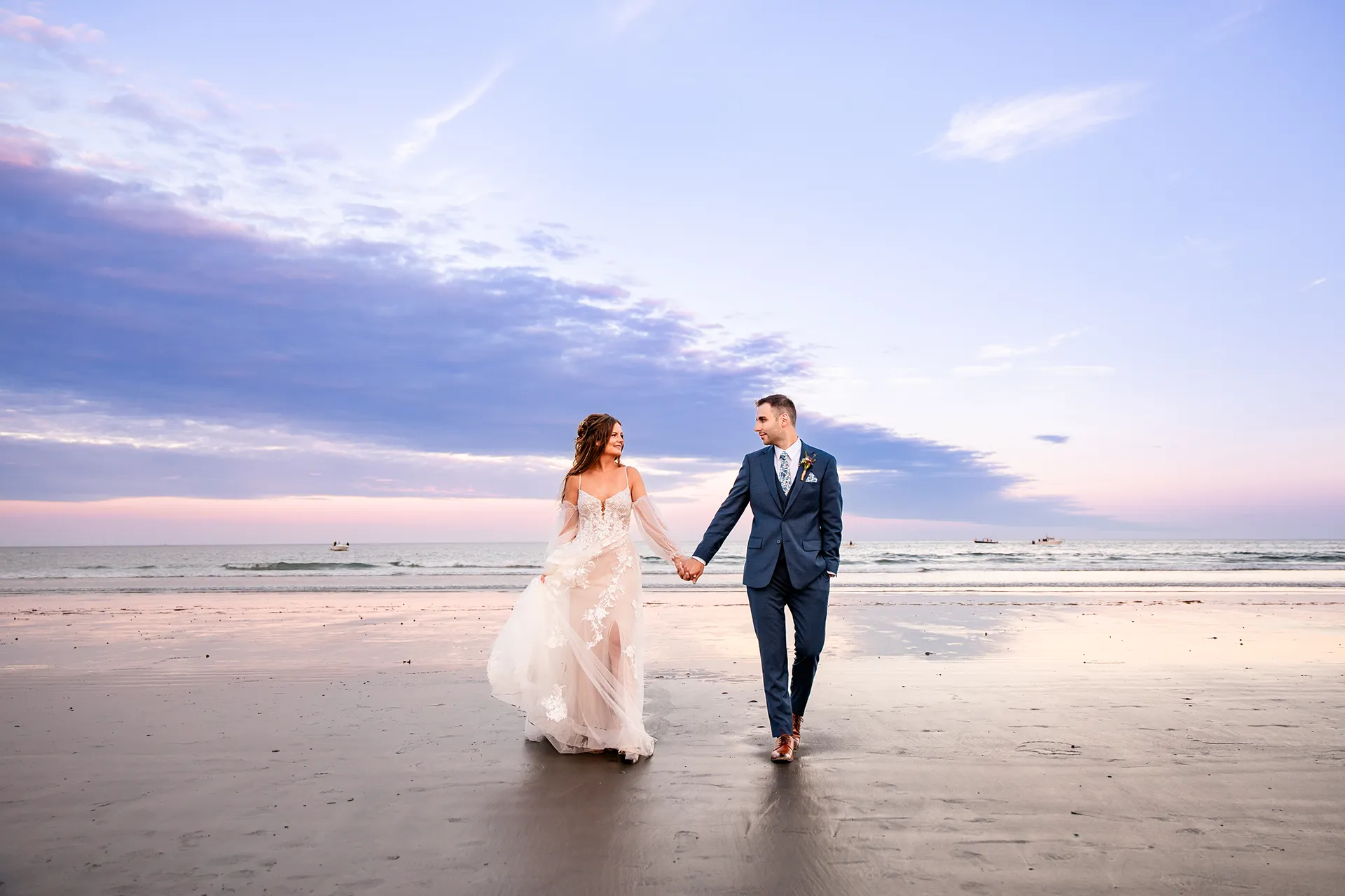 A bride and groom hold hands as they walk on Long Sands Beach during sunset wedding portraits in York, Maine.