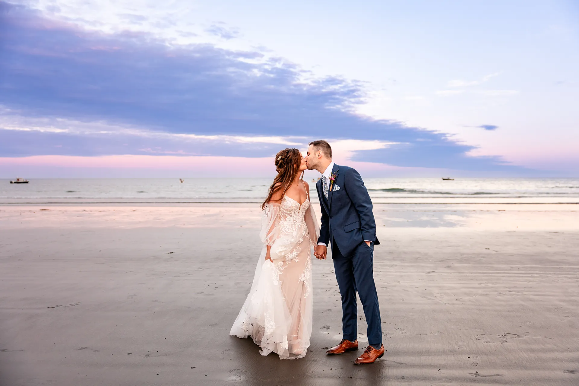 A bride and groom hold hands and kiss as the sun sets during wedding portraits at Long Sands Beach in York, Maine.