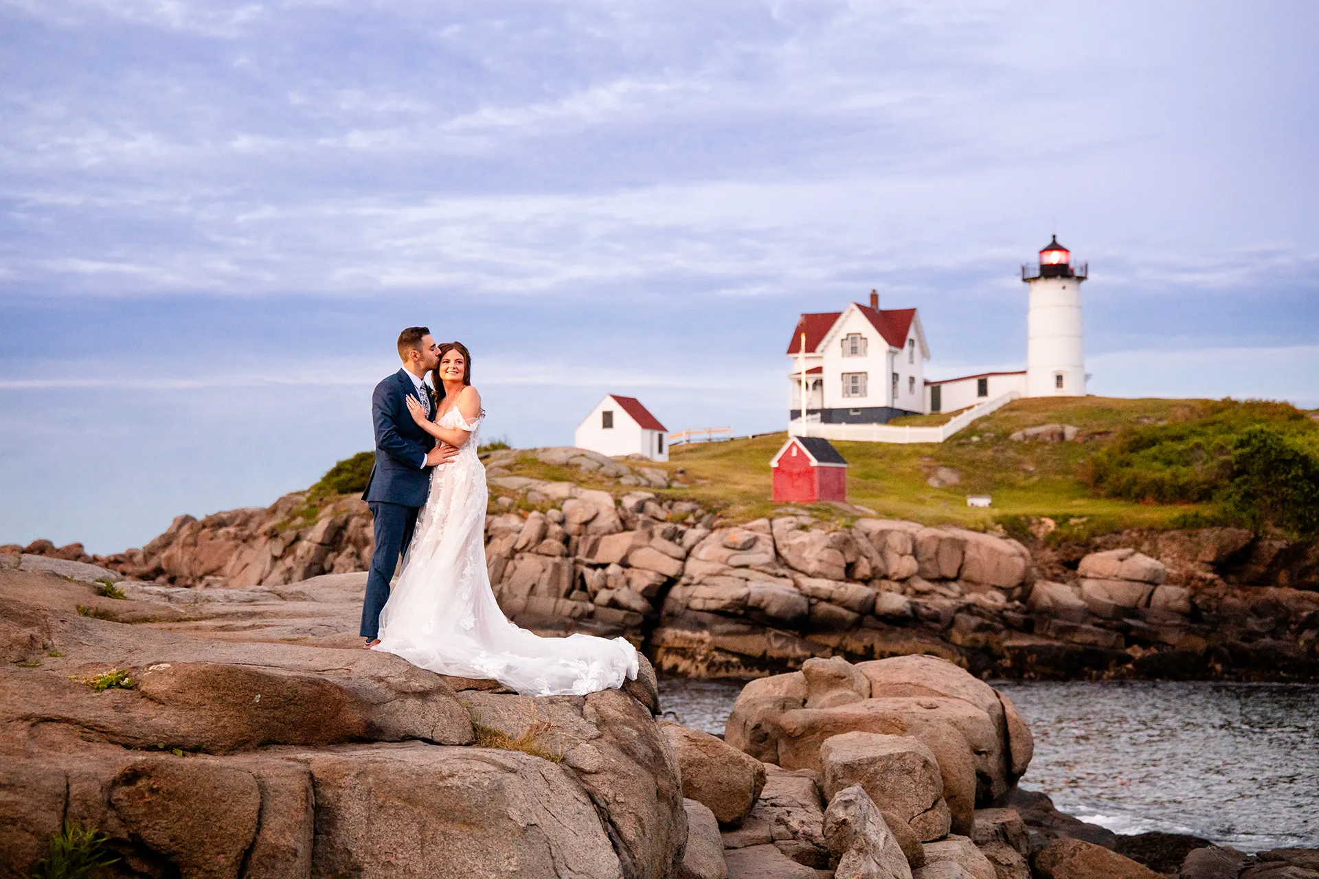 A groom kisses a bride on head during wedding portraits at Nubble Lighthouse in Cape Neddick near York, Maine.