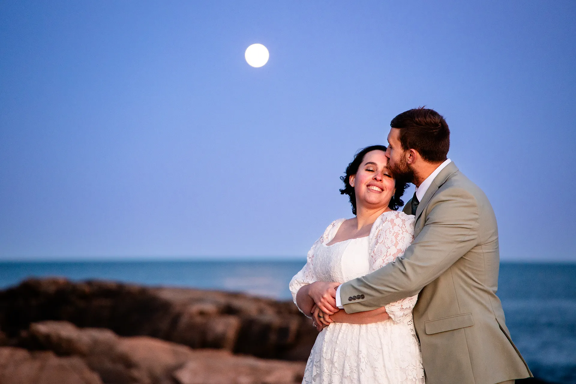 maine wedding photography mystic andrew 34 A groom kisses a bride on the head as the moon shines behind them at Otter Point in Acadia National Park near Bar Harbor, Maine.