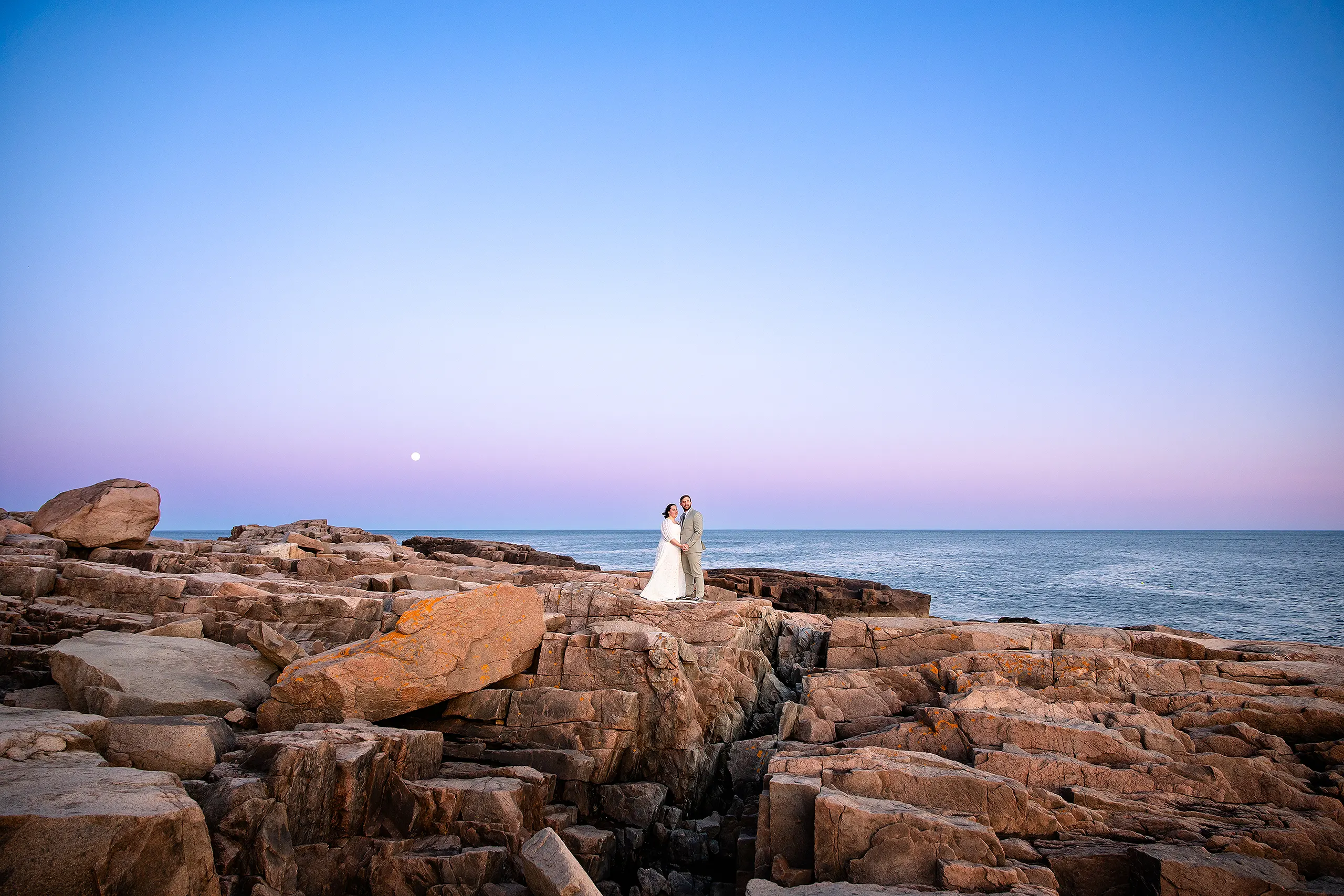 A newly married couple hold hands and pose on a cliff during sunset wedding portraits at Otter Point in Acadia National Park near Bar Harbor, Maine.