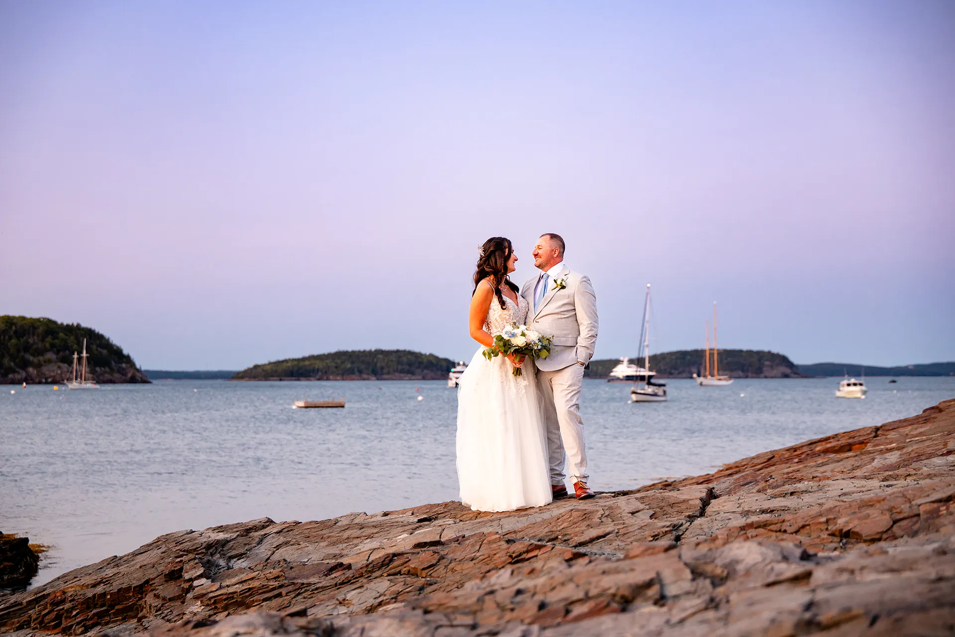 A bride and groom smile at each other on a cliff near the Bar Harbor Inn and Spa during wedding portraits in Bar Harbor, Maine.