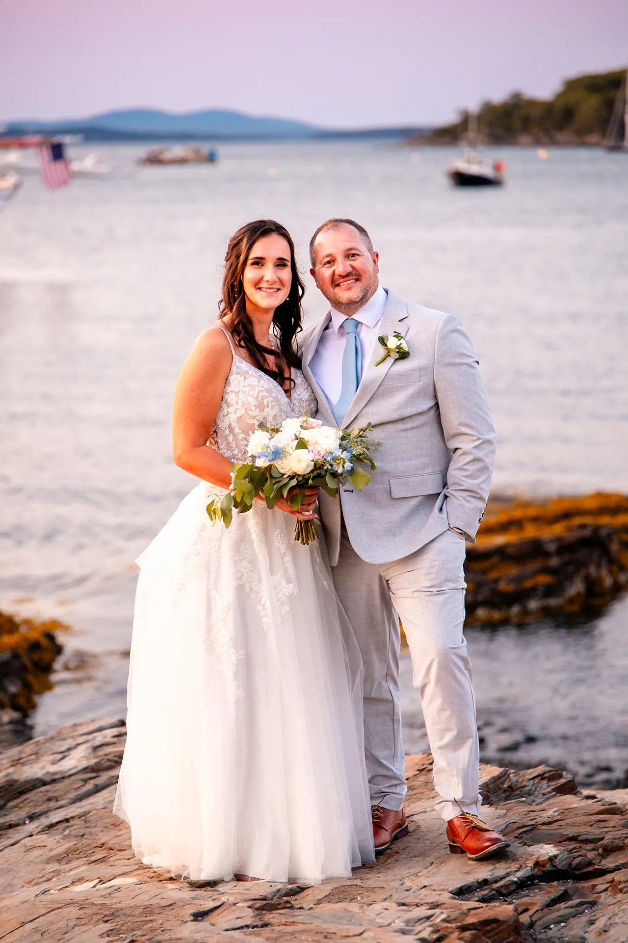 A bride and groom smile and pose overlooking Frenchman Bay near the Bar Harbor Inn and Spa during wedding portraits in Bar Harbor, Maine.