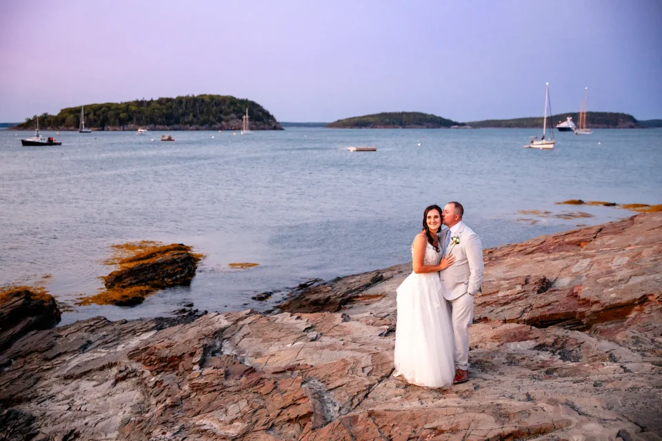 A groom kisses a bride on the cheek during sunset wedding portraits overlooking Frenchman Bay in Bar Harbor, Maine.