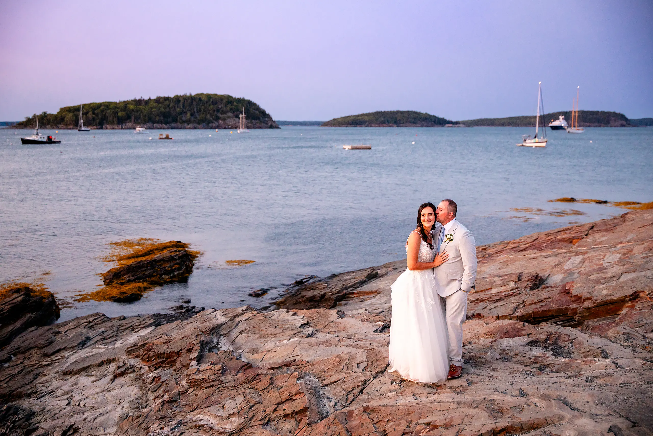 A groom kisses a bride on the cheek during sunset wedding portraits overlooking Frenchman Bay in Bar Harbor, Maine.