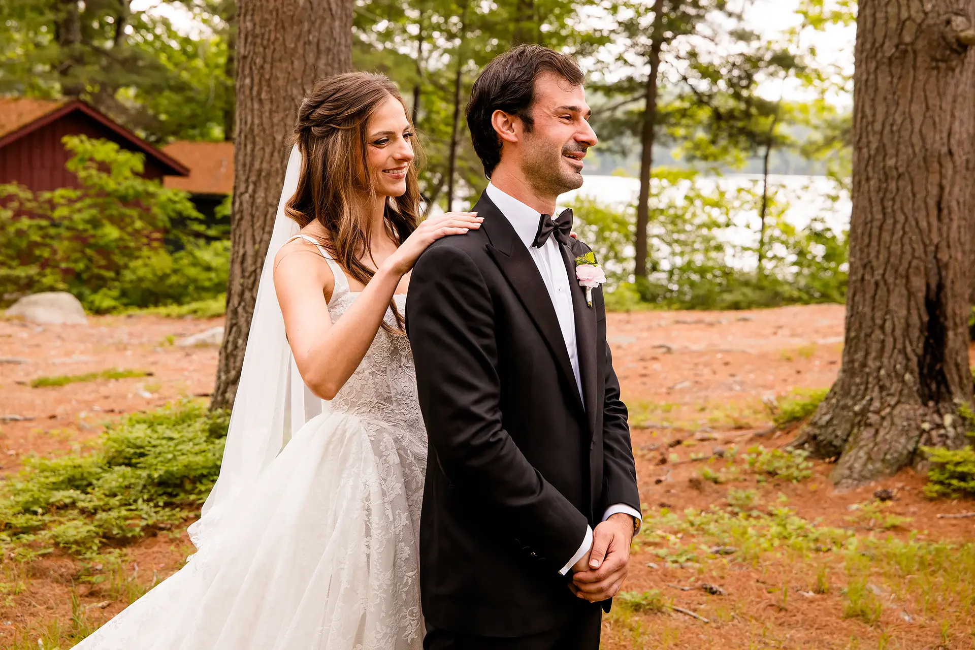 A bride smiles as she rests her hand on a groom's shoulder during a first look at Kingsley Pines Camp in Raymond, Maine.