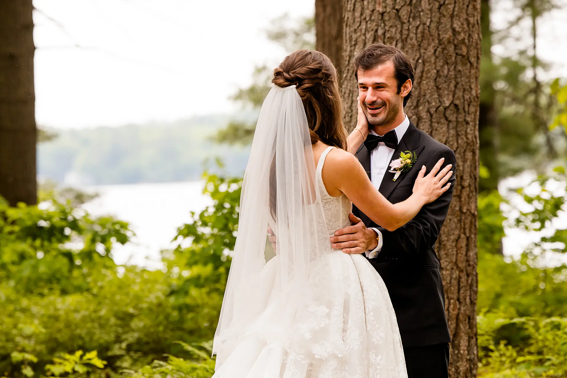 A groom smiles as a bride rests her hand on his face during a first look at Kingsley Pines Camp in Raymond, Maine.