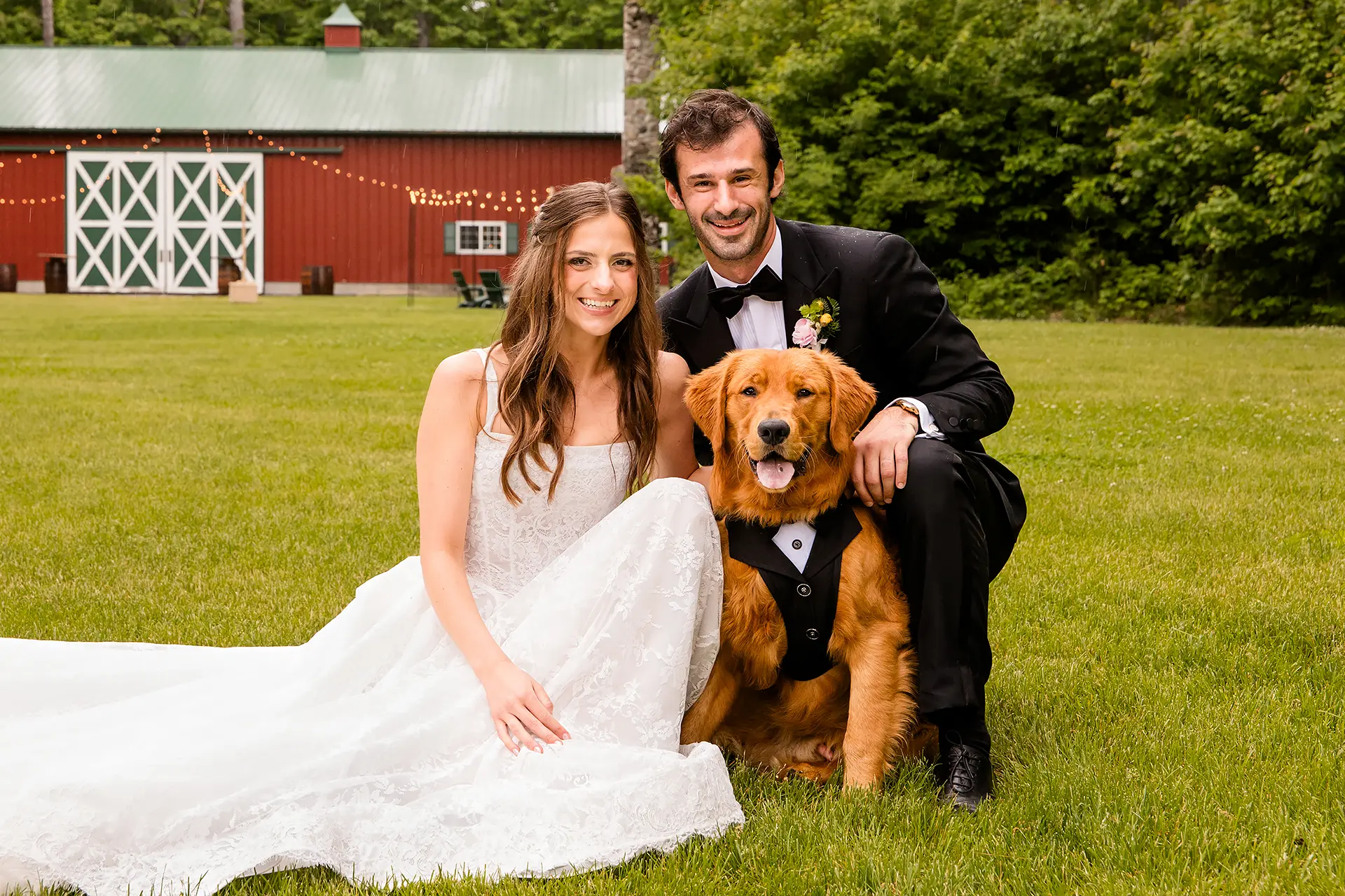 A bride and groom smile and pose with a golden retriever wearing a suit during wedding portraits at Kingsley Pines Camp in Raymond, Maine.