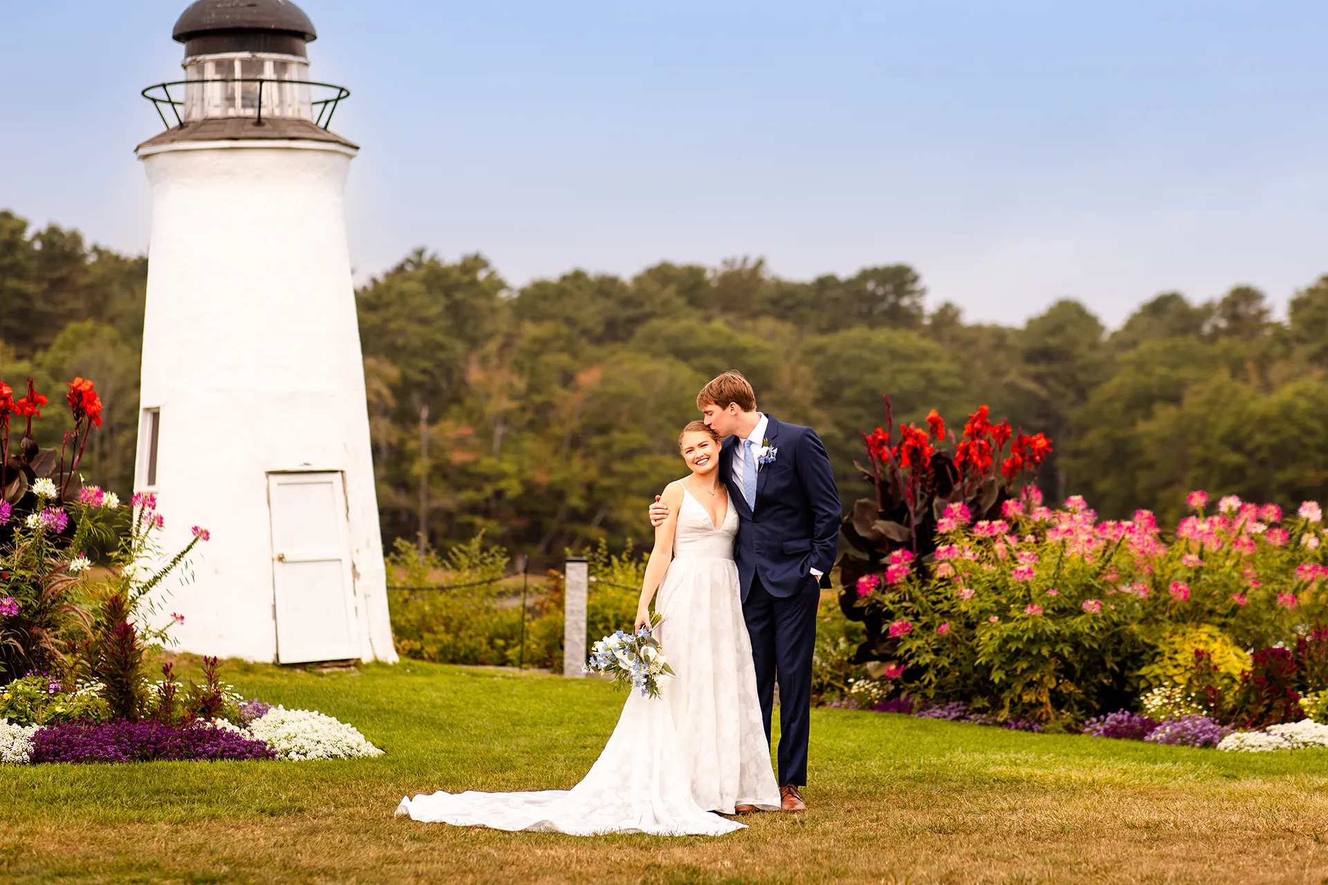 A groom kisses a bride on the head near a lighthouse during wedding portraits at the Nonantum Resort in Kennebunkport, Maine.