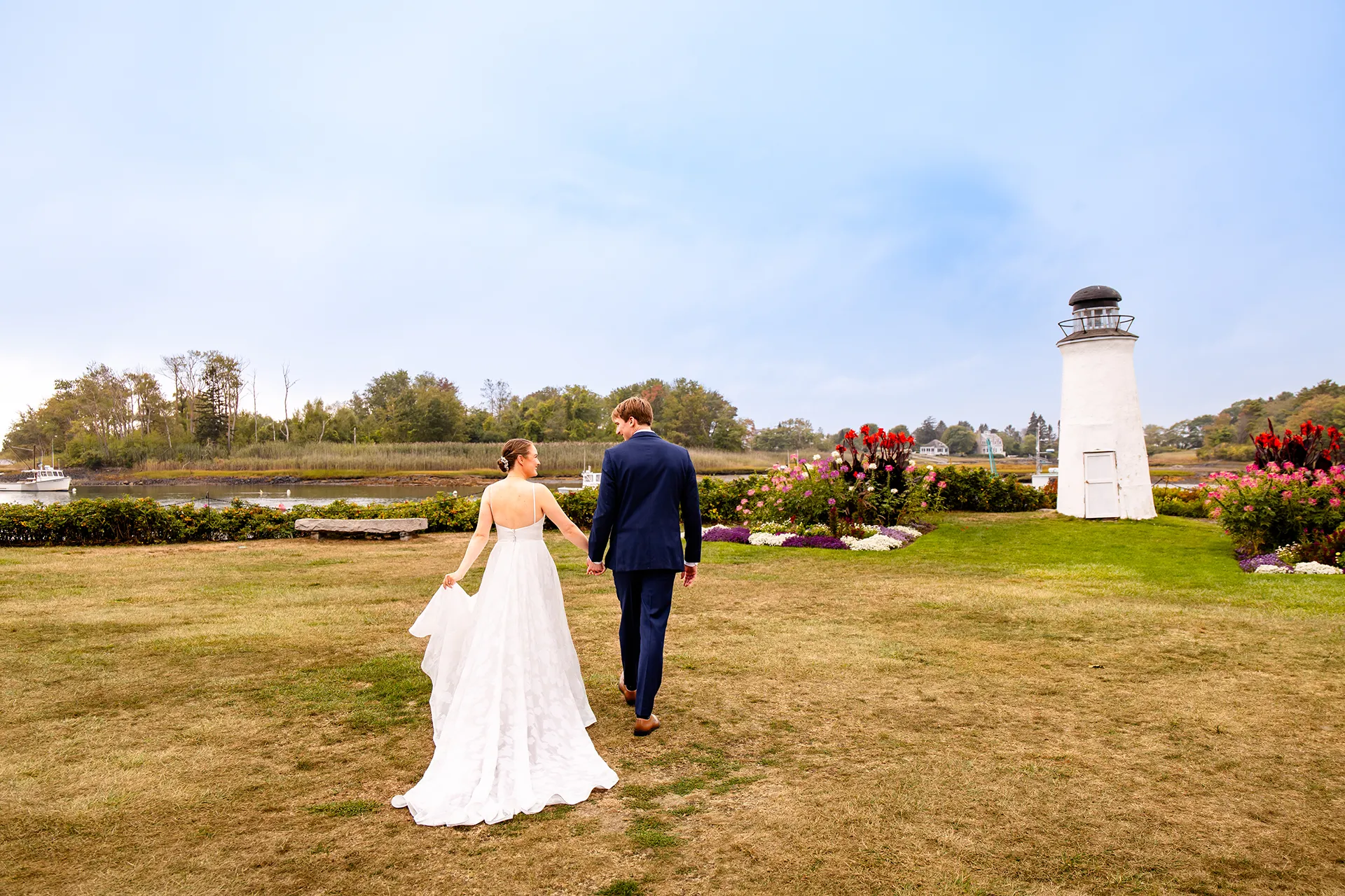 A newly married couple hold hands as they walk toward a lighthouse during wedding portraits at the Nonantum Resort in Kennebunkport, Maine.