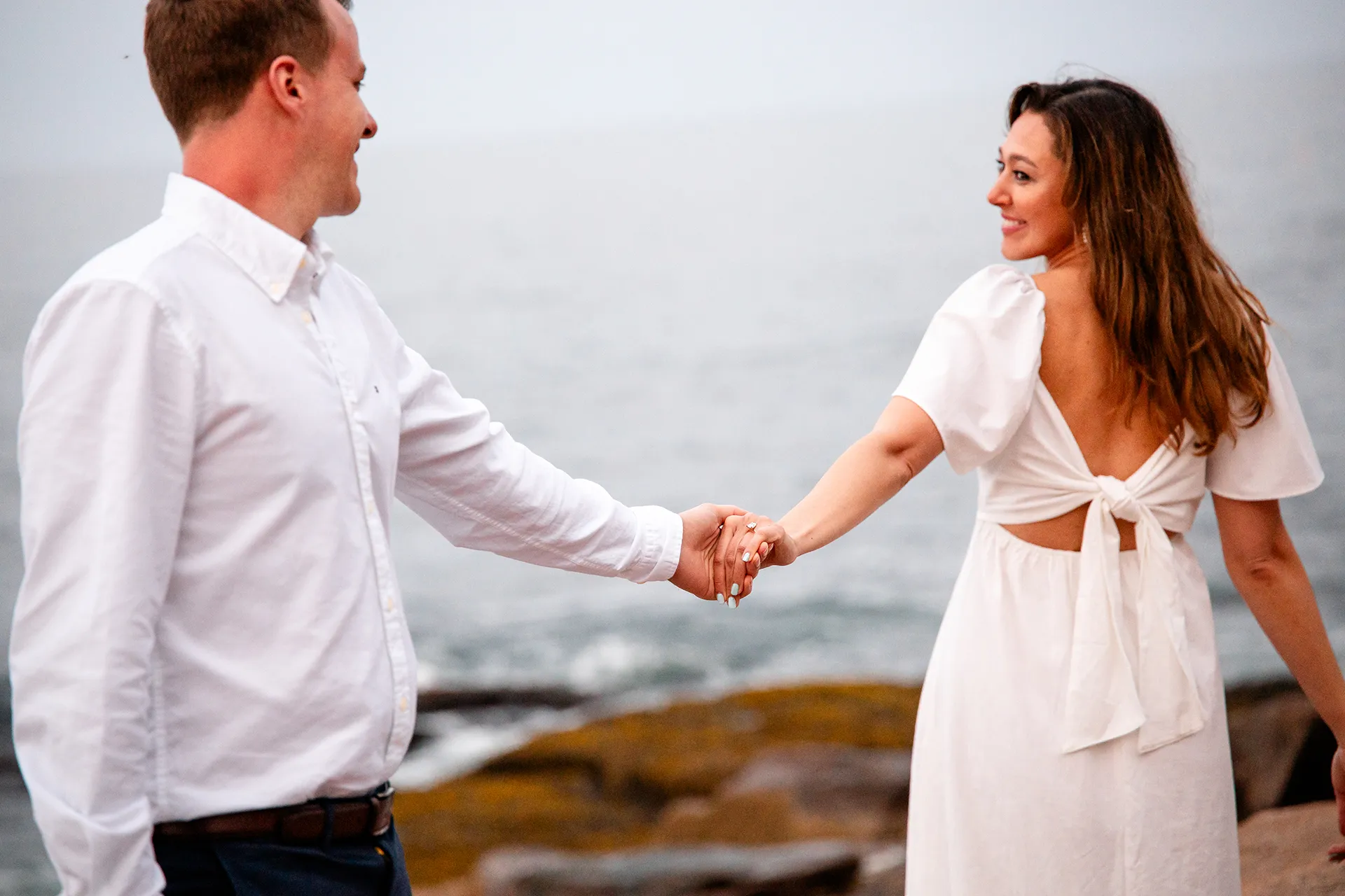 A woman holds a man's hand while looking over her shoulder at him during an engagement session in Acadia National Park, Maine.